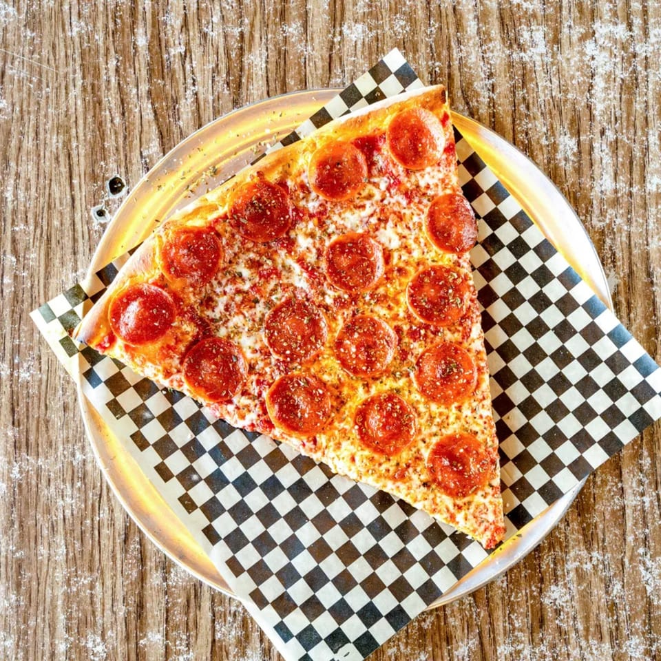 A large slice of pepperoni pizza on checkered paper, served on a metal tray.