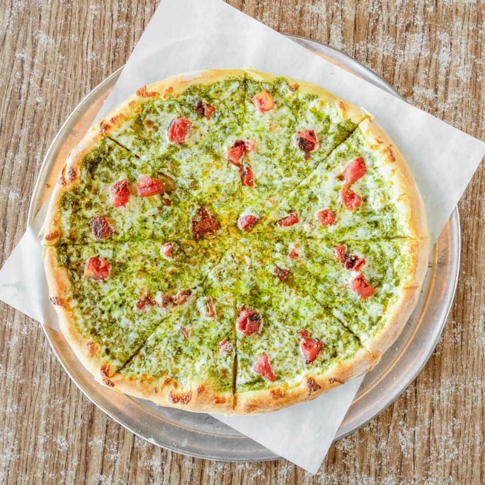 A whole pesto pizza with mozzarella and diced tomatoes on a metal tray, on a wooden table.