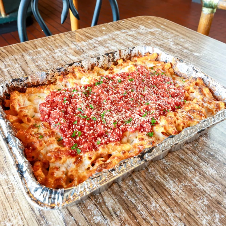 Baked ziti with melted cheese, red sauce, parmesan, and parsley in a foil pan on a wooden table.
