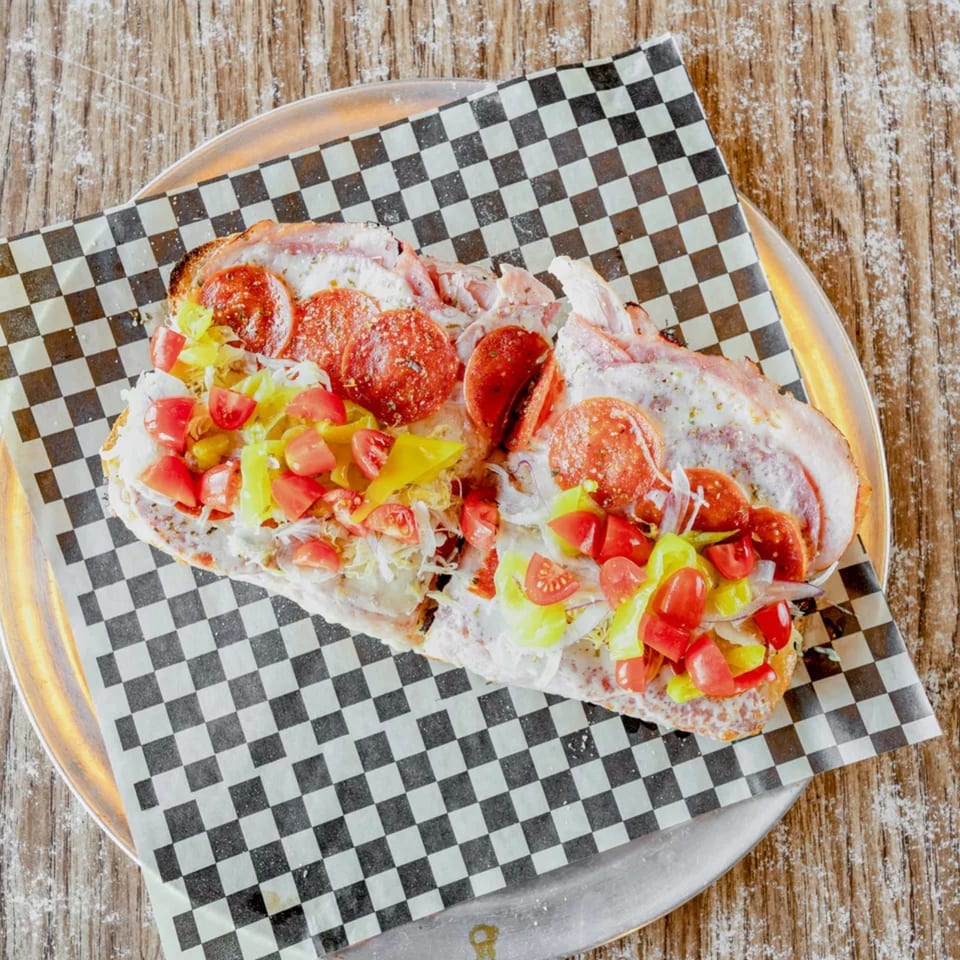A large sub sandwich with pepperoni, tomatoes, and peppers on checkered paper.